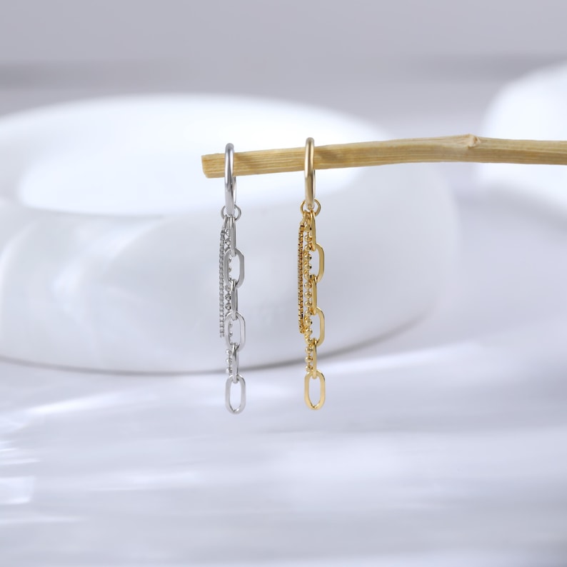 May include: Two dangle earrings, one silver and one gold, suspended from a wooden dowel. The silver earring features a chain design with a row of small, sparkling stones. The gold earring has a similar chain design.