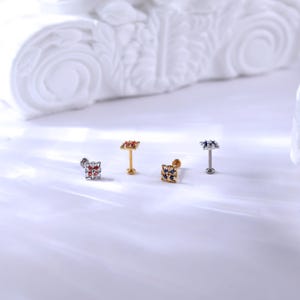 May include: Four different colored square-shaped body jewelry pieces with small gemstones. The jewelry is on a white surface.