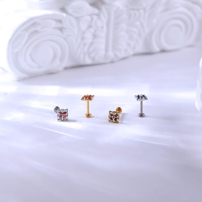 May include: Four different colored square-shaped body jewelry pieces with small gemstones. The jewelry is on a white surface.