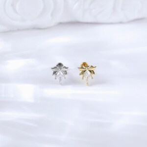 Puede incluir: Dos pendientes de bot&oacute;n, uno plateado y otro dorado, cada uno con un dise&ntilde;o floral y una gema transparente central en forma de l&aacute;grima. Los pendientes est&aacute;n colocados sobre un fondo blanco suave.