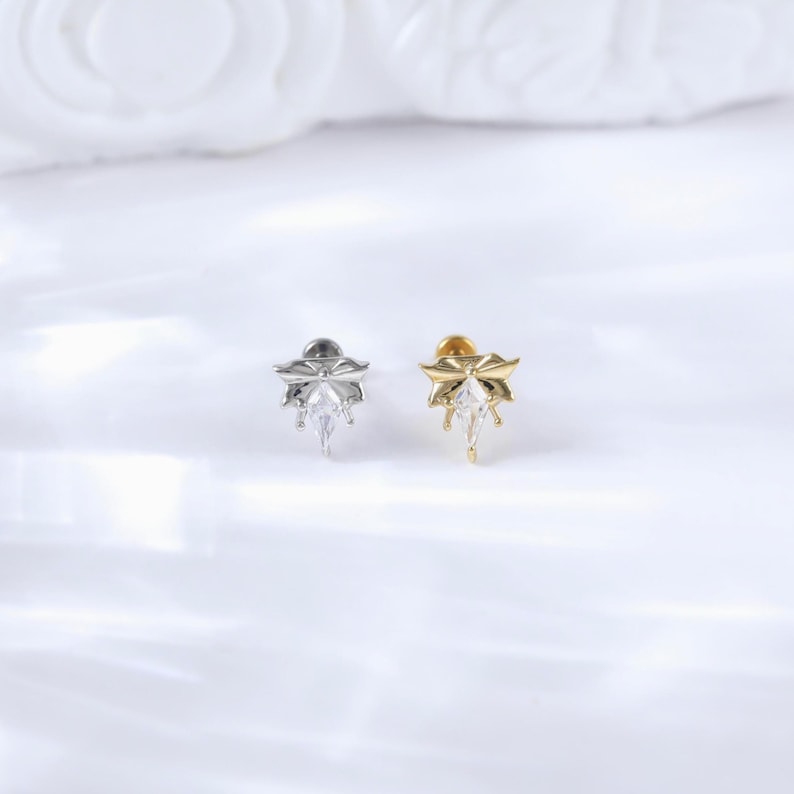 Puede incluir: Dos pendientes de bot&oacute;n, uno plateado y otro dorado, cada uno con un dise&ntilde;o floral y una gema transparente central en forma de l&aacute;grima. Los pendientes est&aacute;n colocados sobre un fondo blanco suave.