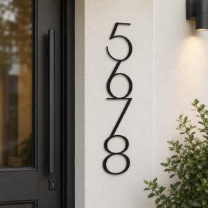 May include: A vertical house number sign featuring the black digits 5, 6, 7, and 8 arranged in a single column. The numbers are constructed with a thin, clean, sans-serif font and are mounted directly onto a light-colored exterior wall surface.