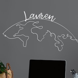 May include: A white wire wall art piece spelling "Lauren" above a world map outline. The art is mounted on a gray wall above a white desk with a laptop, phone, and office supplies. A plant in a woven pot sits on the desk.