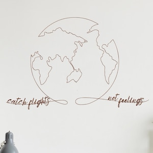 May include: A brown wooden wall art piece featuring a world map outline. The phrase "catch flights not feelings" is written below the map in a cursive font. The artwork is mounted on a white wall.