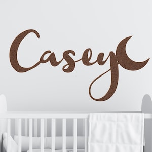 May include: A brown, glittery wall decal featuring the name "Casey" in a cursive script with a crescent moon design. The decal is displayed above a white crib with a white blanket.