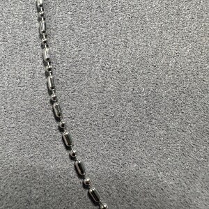 May include: A close-up of a silver-colored ball chain necklace. The chain is composed of small, cylindrical and spherical metal beads. The background is a textured, gray surface.