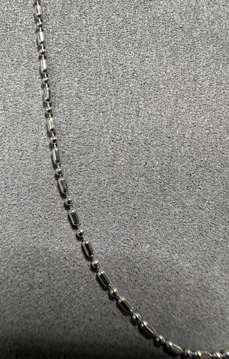 May include: A close-up of a silver-colored ball chain necklace. The chain is composed of small, cylindrical and spherical metal beads. The background is a textured, gray surface.
