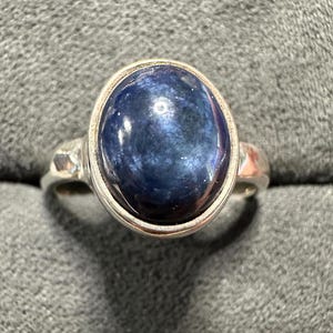 May include: A silver ring featuring a large, oval-shaped, dark blue gemstone. The gemstone has a polished, reflective surface. The ring's band is a simple, classic design, with a hammered texture on the sides.