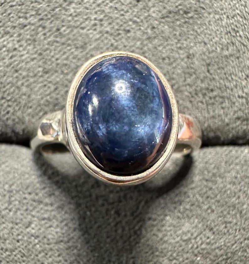 May include: A silver ring featuring a large, oval-shaped, dark blue gemstone. The gemstone has a polished, reflective surface. The ring's band is a simple, classic design, with a hammered texture on the sides.