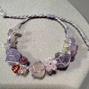 May include: A braided cord bracelet featuring light purple and clear crystal stones, small carved flower-shaped beads in pink and lavender, and a single red teardrop-shaped charm. The stones are secured within the woven light purple cord structure.
