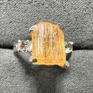 May include: A silver ring featuring a large, rectangular, amber-colored gemstone as the centerpiece. Two smaller, clear gemstones flank the main stone. The ring is set against a gray, textured background.
