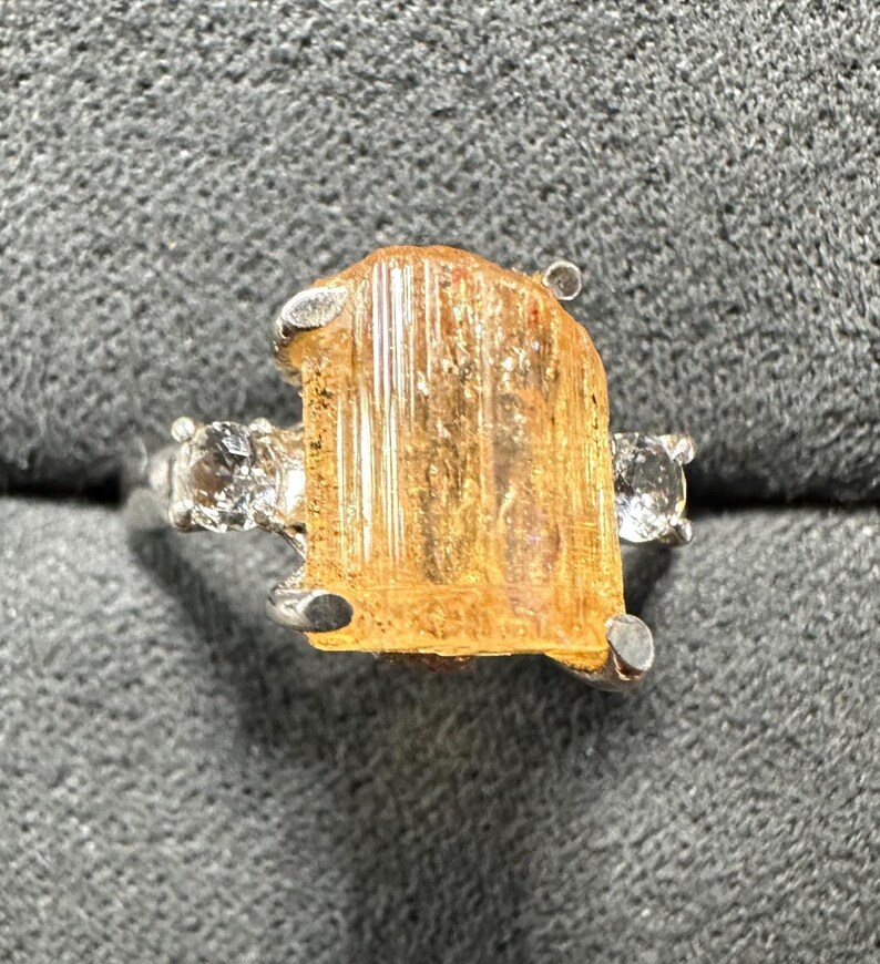May include: A silver ring featuring a large, rectangular, amber-colored gemstone as the centerpiece. Two smaller, clear gemstones flank the main stone. The ring is set against a gray, textured background.