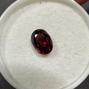 May include: A faceted, oval-shaped, deep red gemstone. The gem is set against a white, textured background within a round, white container. The stone's facets reflect light, creating highlights and shadows.