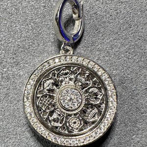 May include: Silver pendant with a circular design. The pendant features a detailed pattern with a central cluster of small, clear stones. The outer edge is lined with additional clear stones, and the bail is a simple, curved design.