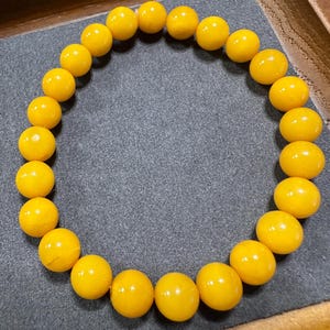 May include: A bracelet featuring a series of spherical, polished yellow beads. The beads are uniform in size and strung together, creating a full circle. The bracelet is presented on a grey surface.