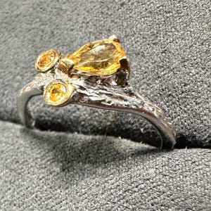 May include: A silver ring with a textured band and three yellow gemstones. The largest gemstone is pear-shaped, while the other two are round. The ring has a unique, organic design.