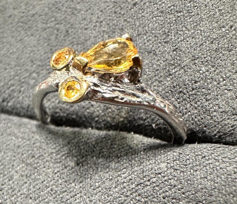 May include: A silver ring with a textured band and three yellow gemstones. The largest gemstone is pear-shaped, while the other two are round. The ring has a unique, organic design.