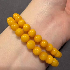 May include: Two amber-coloured beaded bracelets. Each bracelet is made of round, polished beads. The bracelets are worn together, resting on a person's wrist. The beads have a warm, golden-yellow hue, and the bracelets appear to be made of a natural material.