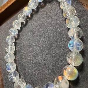 Pode incluir: Uma pulseira de contas transparentes e iridescentes. As contas redondas t&ecirc;m uma qualidade transl&uacute;cida, com flashes de cores do arco-&iacute;ris. A pulseira &eacute; exibida em uma superf&iacute;cie cinza escuro, dentro de uma bandeja de madeira.