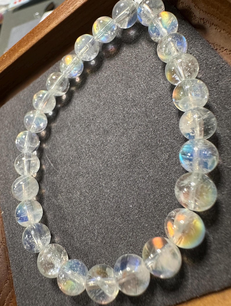 Pode incluir: Uma pulseira de contas transparentes e iridescentes. As contas redondas t&ecirc;m uma qualidade transl&uacute;cida, com flashes de cores do arco-&iacute;ris. A pulseira &eacute; exibida em uma superf&iacute;cie cinza escuro, dentro de uma bandeja de madeira.