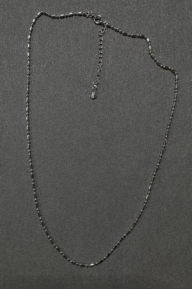 May include: A delicate silver necklace with a beaded design. The necklace features small, cylindrical beads strung together to form a chain. The clasp and extender chain are visible, and the necklace is displayed against a dark gray background.