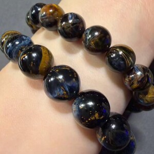 May include: Two bracelets with large, round beads. The beads are a mix of dark blue, black, and gold, with a marbled appearance. The bracelets are worn on a wrist, showcasing the jewelry's colors and textures.