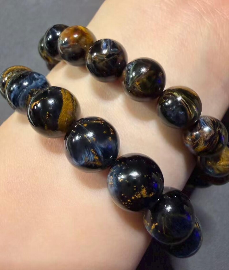 May include: Two bracelets with large, round beads. The beads are a mix of dark blue, black, and gold, with a marbled appearance. The bracelets are worn on a wrist, showcasing the jewelry's colors and textures.
