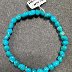 May include: A turquoise bracelet made of smooth, rounded beads. The bracelet is strung on a clear elastic cord. The tag attached to the bracelet reads "Kingman Turquoise".