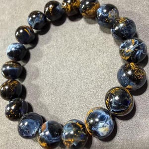 May include: A beaded bracelet featuring polished, round beads. The beads are primarily dark blue with swirling patterns and flecks of gold and yellow. The bracelet is displayed on a gray surface.