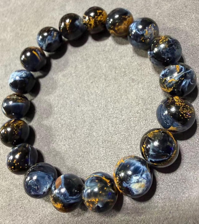 May include: A beaded bracelet featuring polished, round beads. The beads are primarily dark blue with swirling patterns and flecks of gold and yellow. The bracelet is displayed on a gray surface.