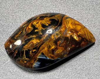 May include: A polished, oval-shaped gemstone with a swirling pattern of gold, black, and blue hues. The stone has a glossy surface, reflecting light and showcasing the natural variations in color and texture. The gemstone is a unique, eye-catching piece.