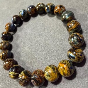 May include: A bracelet made of round, polished beads. The beads are a mix of brown, gold, and blue, with a swirling pattern. The bracelet is resting on a gray surface.