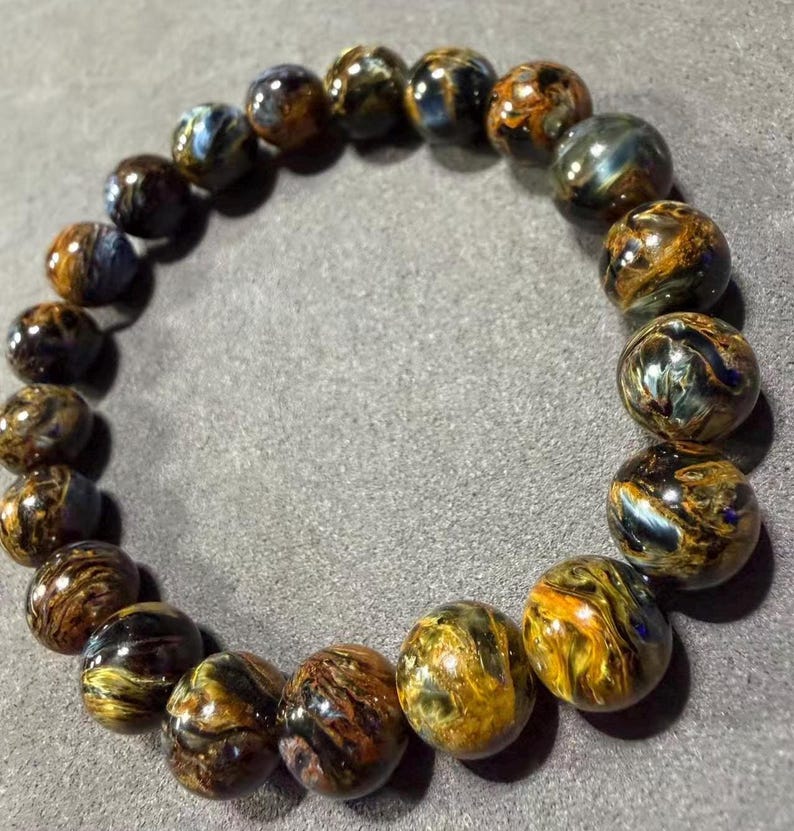 May include: A bracelet made of round, polished beads. The beads are a mix of brown, gold, and blue, with a swirling pattern. The bracelet is resting on a gray surface.