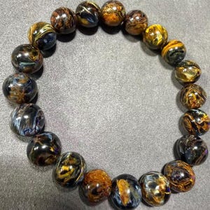 May include: A beaded bracelet featuring polished, round gemstones in shades of brown, gold, and blue. The bracelet is displayed on a gray surface, showcasing the natural patterns and colors of the stones. The beads are approximately 0.5 inches in diameter.