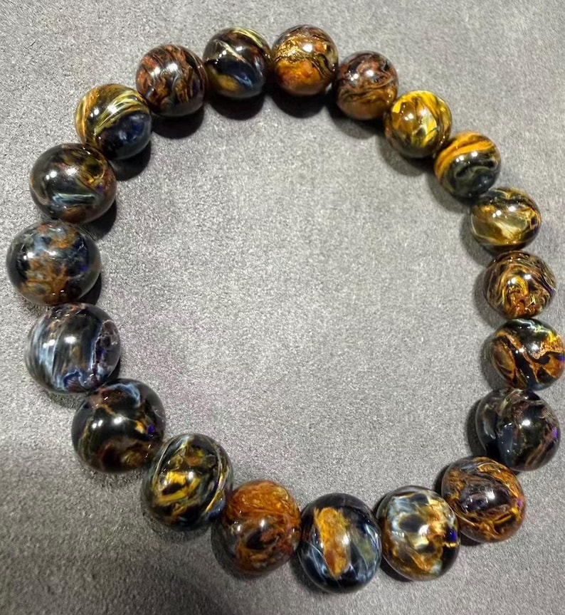 May include: A beaded bracelet featuring polished, round gemstones in shades of brown, gold, and blue. The bracelet is displayed on a gray surface, showcasing the natural patterns and colors of the stones. The beads are approximately 0.5 inches in diameter.