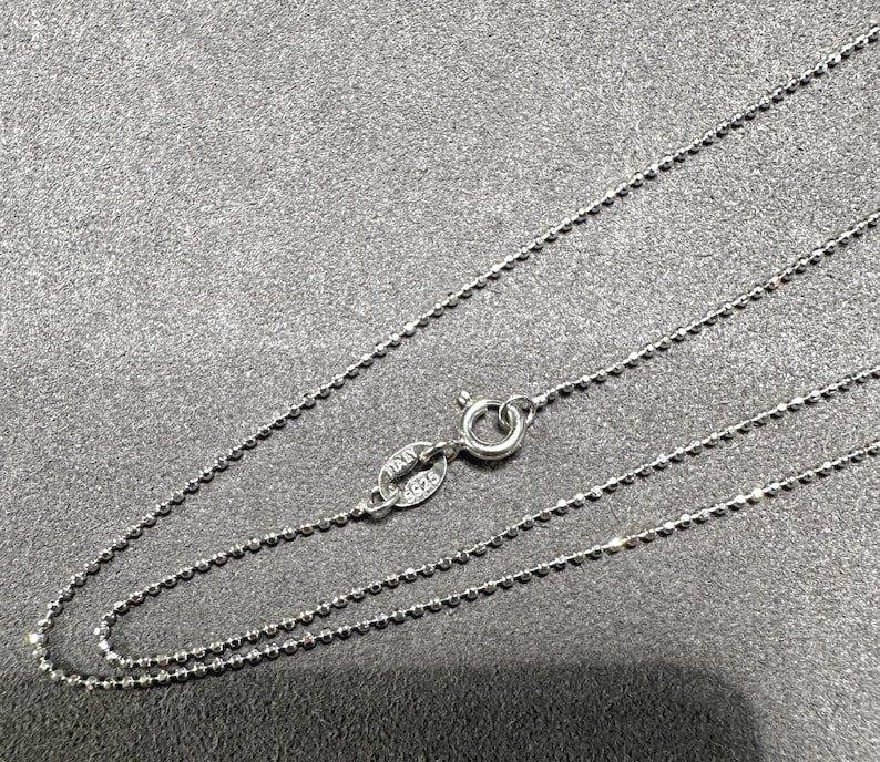 May include: A delicate silver necklace with a ball chain design. The necklace features a small clasp and a tag that reads "ITALY 925". The chain is displayed on a gray surface.