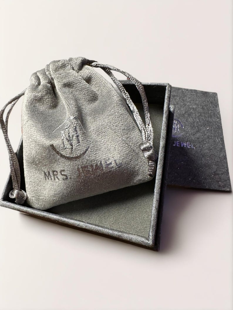May include: A gray velvet jewelry bag with a drawstring closure sits inside a matching gray box. The bag is embossed with the words "MRS. JEWEL" and a logo. A gray card with the logo and the word "JEWEL" is also visible.