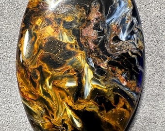 May include: Oval-shaped Pietersite gemstone with a polished surface. The stone features a swirling pattern of gold, black, blue, and brown colors. The gemstone is set against a gray background.