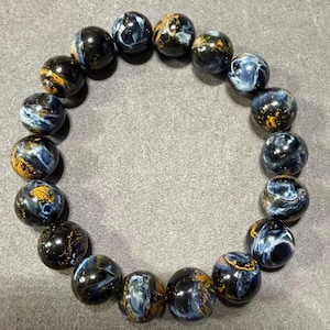 May include: A beaded bracelet featuring round, polished beads. The beads are primarily dark blue with swirling patterns of lighter blue and gold. The bracelet is displayed on a gray surface.