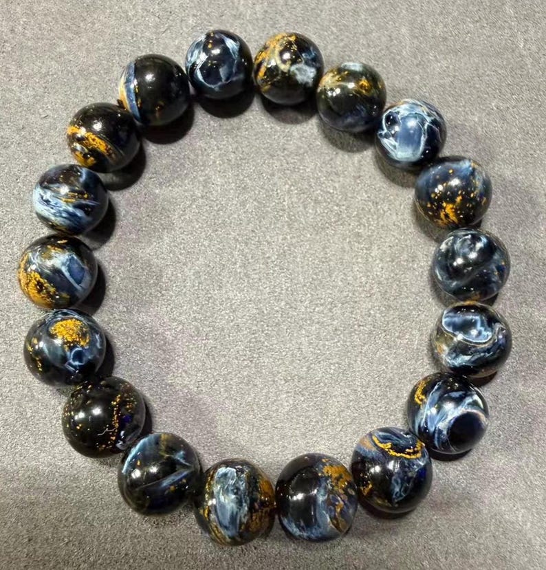May include: A beaded bracelet featuring round, polished beads. The beads are primarily dark blue with swirling patterns of lighter blue and gold. The bracelet is displayed on a gray surface.