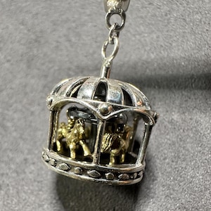 May include: Silver-toned carousel charm pendant with gold-colored horses inside a cage-like structure. The charm is suspended from a silver chain, showcasing a detailed miniature design. The carousel horses are in a running position.