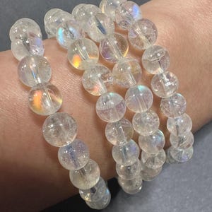 Pode incluir: Quatro pulseiras de contas claras e iridescentes. Cada pulseira &eacute; feita de contas redondas e transl&uacute;cidas que refletem um arco-&iacute;ris de cores. As pulseiras s&atilde;o empilhadas, criando um efeito em camadas. As contas parecem ser feitas de um material semelhante ao cristal.