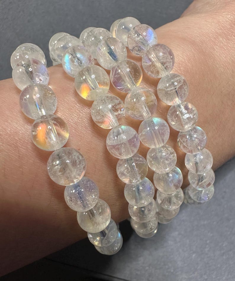 Pode incluir: Quatro pulseiras de contas claras e iridescentes. Cada pulseira &eacute; feita de contas redondas e transl&uacute;cidas que refletem um arco-&iacute;ris de cores. As pulseiras s&atilde;o empilhadas, criando um efeito em camadas. As contas parecem ser feitas de um material semelhante ao cristal.