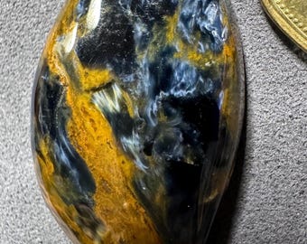 Könnte beinhalten: Ein polierter, tropfenförmiger Edelstein mit einem wirbelnden Muster aus blauen, schwarzen und goldbraunen Farbtönen. Die Oberfläche des Steins ist glänzend, reflektiert das Licht und zeigt seine natürlichen Farben. Eine Goldmünze ist teilweise sichtbar.