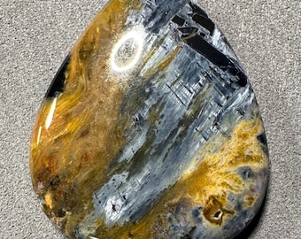 May include: A polished, teardrop-shaped gemstone with a mix of colors. The stone features swirling patterns of blue, gold, black, and brown. The surface is smooth and reflective, with a glossy finish. The gemstone is likely used for jewelry or display.