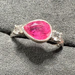 May include: A silver ring featuring a pear-shaped, vibrant pink gemstone. The gemstone is the focal point, set in a silver bezel. Two smaller, clear gemstones flank the main stone, adding sparkle. The ring is set against a gray background.
