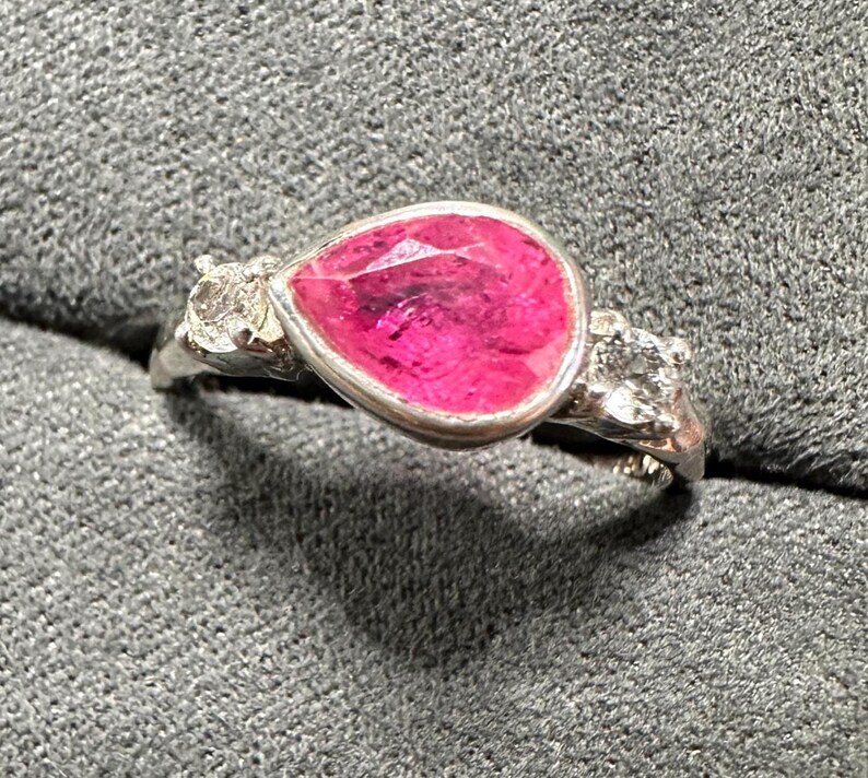 May include: A silver ring featuring a pear-shaped, vibrant pink gemstone. The gemstone is the focal point, set in a silver bezel. Two smaller, clear gemstones flank the main stone, adding sparkle. The ring is set against a gray background.
