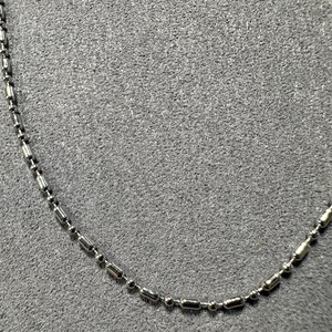 May include: A silver-colored necklace with a ball and tube chain design. The necklace is made of small, shiny metal beads and cylindrical links. The chain is displayed against a gray background.