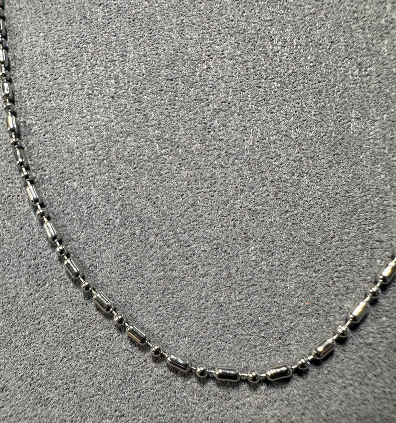 May include: A silver-colored necklace with a ball and tube chain design. The necklace is made of small, shiny metal beads and cylindrical links. The chain is displayed against a gray background.