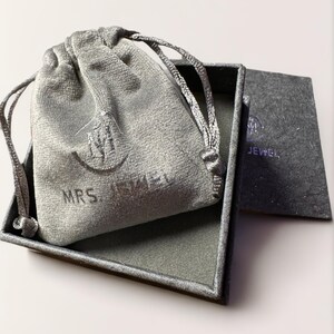 May include: A gray velvet jewelry pouch with the text "MRS. JEWEL" on it, inside a gray square box with a gray velvet lining. The box has a gray paper insert with the text "MRS. JEWEL" on it.